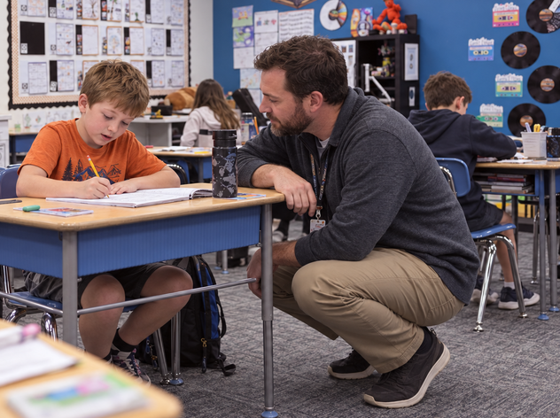 Teacher kneeling beside a student to help with written work.