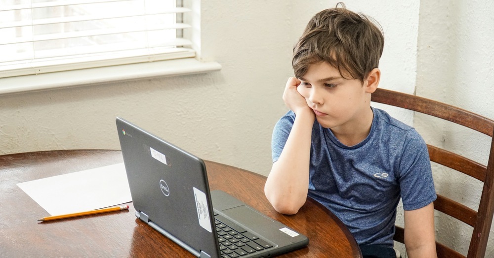 A boy sits at a wooden table, chin resting in his hand, staring blankly at an open Chromebook.