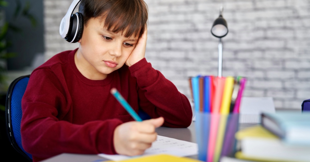 A child wearing white headphones sits at a desk with a pencil in hand, looking down at a notebook with a tired, distracted expression.